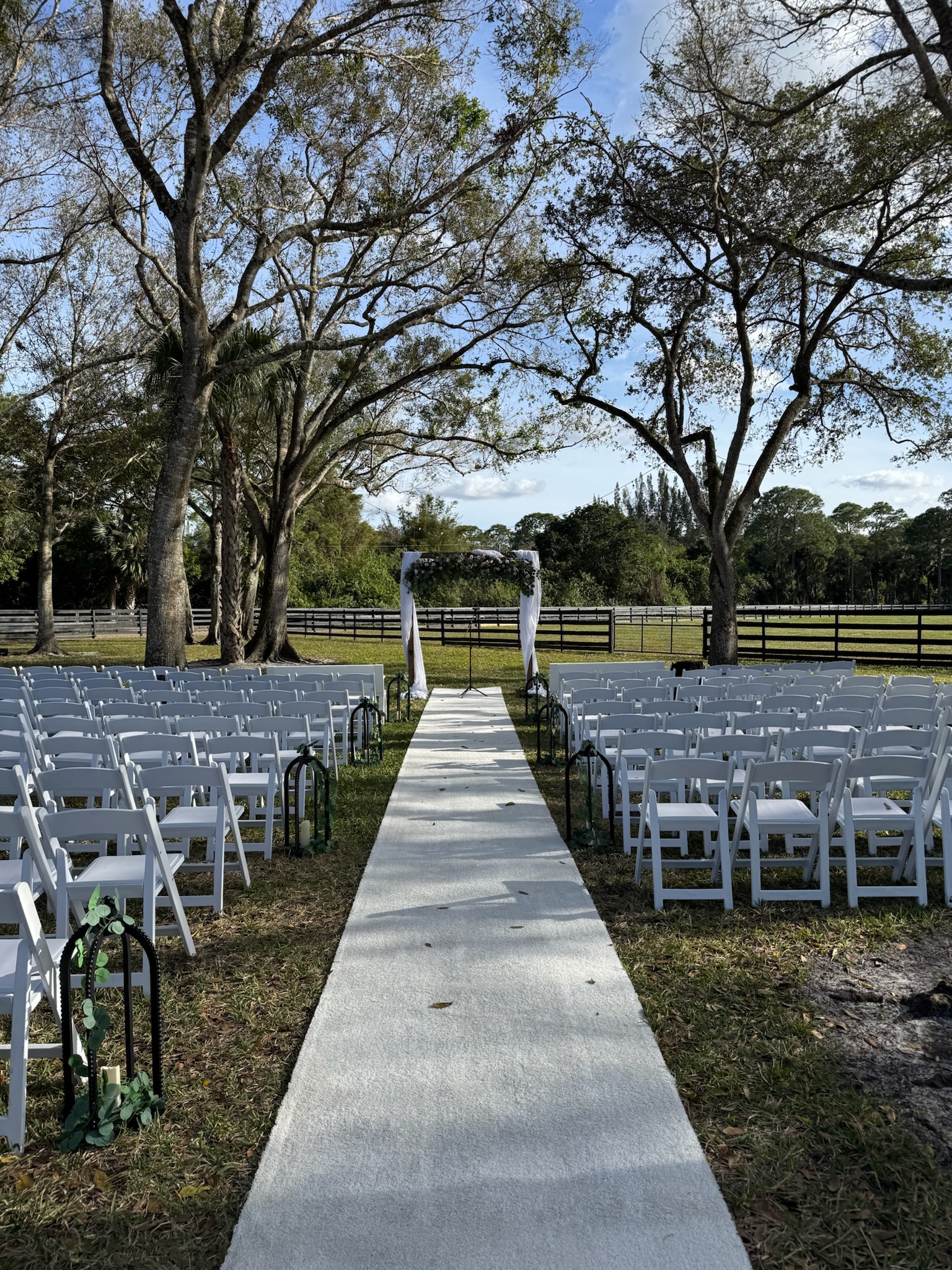 Romantic outdoor ceremony setup at a golf course with white chairs, floral arch, and a lake backdrop at sunset